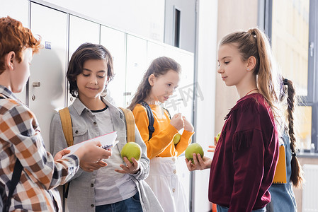 小学生们拿着新鲜苹果站在学校大厅里的朋友们身边，手里拿着笔记本和背包 