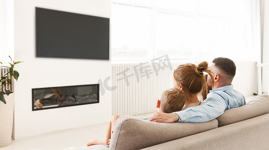 年轻的家庭、父亲、母亲和两个小孩坐在壁炉前的沙发上看电视，在家里聚精会神地享受闲暇时光的背景图