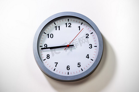 grey摄影照片_Grey Wall Clock show the time. Modern wall clock with grey frame on white background. Close up to a clock, with black time pointer and red second pointer. Shadow from the daylight.  