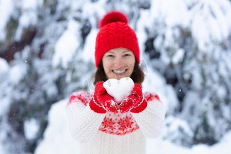 冬天穿着针织毛衣的年轻女子拿着心形雪球.女孩在家庭雪地格斗游戏。身穿针织手工制作的帽子和带圣诞雪花图案的手套的女性，在雪地公园玩乐