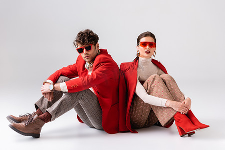 穿着红色夹克和太阳镜的时髦夫妇坐在地板上，摆出一副灰色的样子