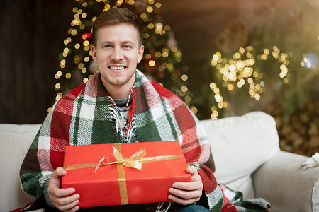 年轻英俊的男人穿着暖和的毛衣，拿着礼物坐在沙发上，房间里装饰着庆祝新年和圣诞节的气氛