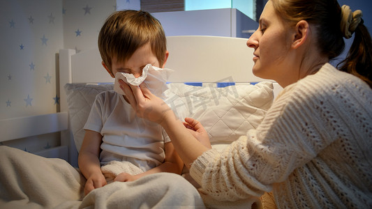 说话的小男孩摄影照片_生病的小男孩用纸巾捂住鼻子，在晚上睡觉前和妈妈说话。儿童疾病、疾病和父母照料的概念