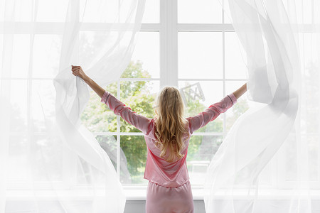 粉色窗帘摄影照片_穿着粉色睡衣的金发年轻女子醒来，看着卧室里的大玻璃窗
