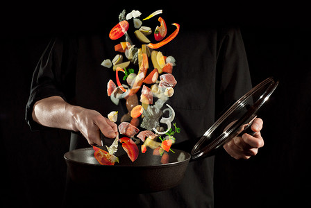 胡萝卜土豆摄影照片_穿着黑色衬衫的男人在深色背景下准备食物.肉、土豆、西红柿、胡萝卜和其他蔬菜在空气中飘扬.模糊的艺术效果。运动、飞行和悬浮概念.