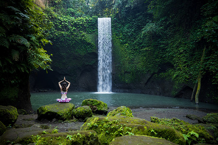 瑜伽莲花姿势。年轻的白人妇女坐在石头上，沉思着练习瑜伽，在瀑布边弹跳。手举起来在namaste mudra 。瑜伽撤退。从后面看蒂布马纳瀑布巴厘