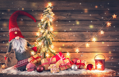 一棵摄影照片_圣诞节背景。圣诞老人或侏儒拿着一棵冷杉树, 上面有圣诞灯, 周围都是礼品盒和乡村木上发光的灯笼