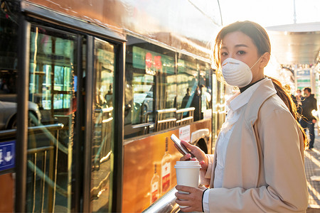 戴口罩女摄影照片_戴口罩的年轻女人站在公交车站