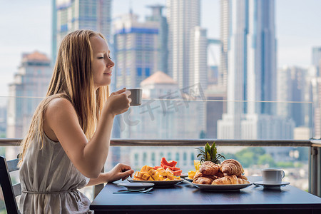 一个年轻的女人正在阳台上吃早饭。早餐桌与咖啡果子和面包 croisant 在阳台反对大都市的背景
