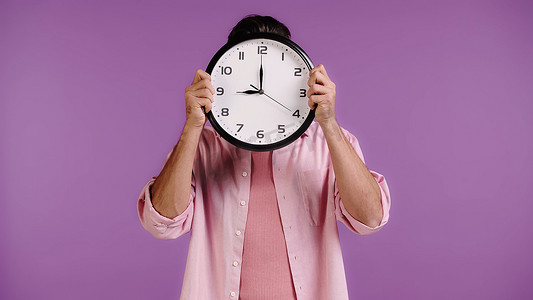 young man in pink shirt obscuring face with clock isolated on purple