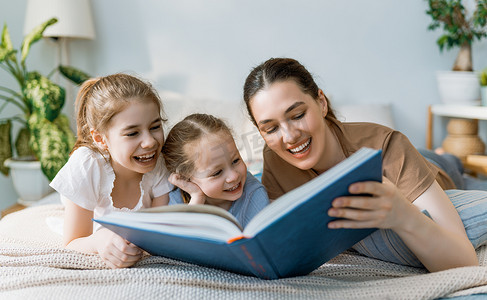 快乐温馨的家庭。漂亮的年轻母亲在家里的床上给女儿们读书.