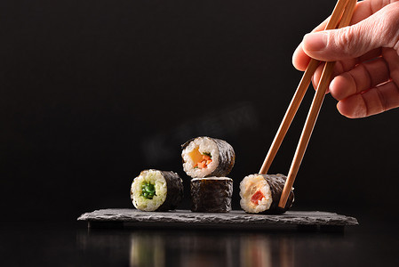 背景手在黑色背景的深色木桌上，用筷子在板子上采摘4个不同种类的蔬菜寿司。前面的景色1.横向构成.
