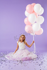 漂亮的小女孩穿着公主礼服坐在孤立在紫罗兰色的背景, 拿着一堆气球