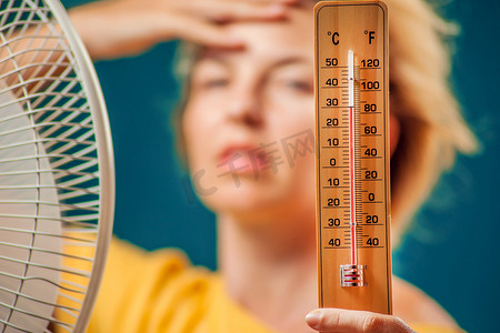 炎热温度计摄影照片_一个女人的肖像在风扇前遭受的热保持温度计。炎热天气概念