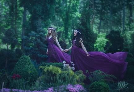 紫色公主摄影照片_两个公主穿着紫色的连衣裙在风中挥舞着, 在一个美丽的花园的背景下行走。两个不同的姐妹是金发和黑发。童话夏天