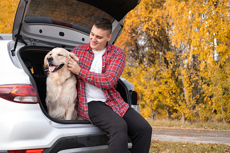 宠物主人摄影照片_秋天的森林里，一个男人和他的狗坐在汽车后备箱里。和宠物一起旅行一个关于黄金猎犬的本质和主人的旅行.