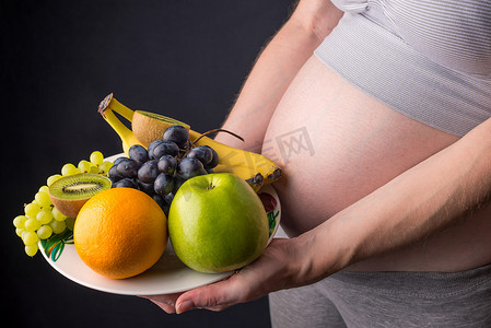 腹部水肿摄影照片_孕妇腹部拿着盘子里的水果。为控制体重和怀孕期间的健康饮食概念