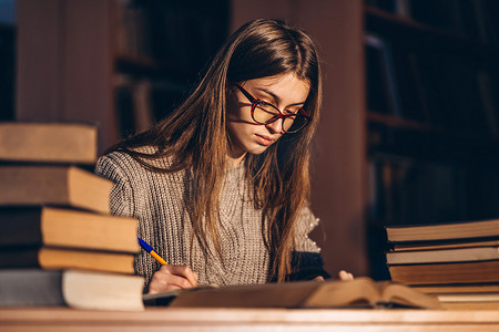 戴眼镜的年轻学生在准备考试.晚上的女孩拿着一堆书坐在图书馆的桌子旁.