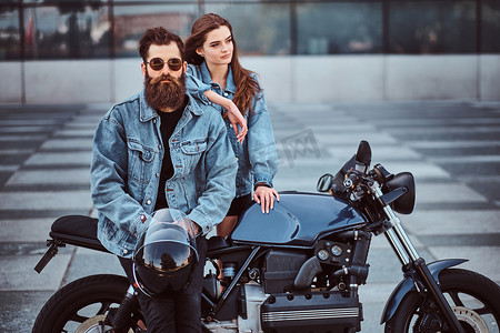 迷人的时髦夫妇-胡子的野蛮男性在太阳镜和牛仔裤夹克坐在复古摩托车和他年轻的感性女孩站在附近, 摆在一个摩天大楼.