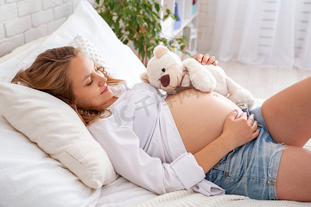 躺着休息摄影照片_孕妇躺在床上躺着。摸她光溜溜的肚子准妈妈在舒适明亮的房间里。和未出生的宝宝休息和交流。等待的概念。分娩前准备.