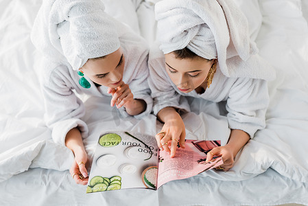 穿着浴袍、耳环的时尚女性的头顶视图，头上有毛巾，躺在床上阅读杂志