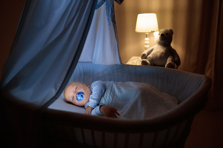 可爱的宝宝与奶嘴晚上睡在蓝色摇篮与冠层。小男孩穿着睡衣小睡片刻在黑暗的房间里，与婴儿床、 灯和玩具熊。孩子的睡觉时间。卧室和托儿所室内.