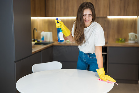年轻漂亮的女人，戴着黄色手套，手里拿着洗涤剂喷雾，用抹布擦厨房桌上的灰尘