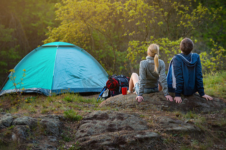 两个徒步旅行者带着背包在夕阳下温暖的阳光下, 在帐篷附近休息。旅行, 假期, 假期和冒险概念。森林山风景背景