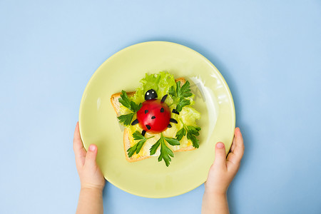 创意瓢虫摄影照片_给孩子们的美食艺术。孩子们手拿着装有瓢虫三明治蓝色背景的盘子。如何在家里为孩子做有创意的早餐。一步一步地说明，从上往下看