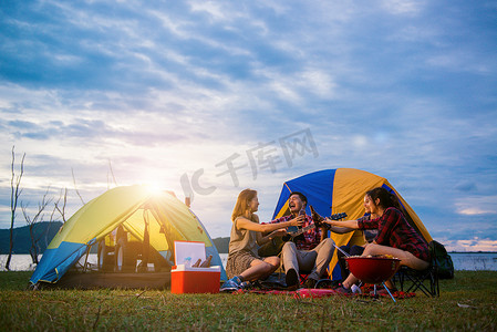 露营躺平摄影照片_组的男人和女人享受露营野餐和烧烤在帐篷中背景的湖。年轻混合种族亚洲女人和男人。年轻人们手里烘培和欢呼瓶啤酒.