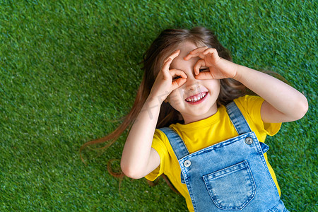 穿着牛仔连衣裙的快乐的小女孩躺在绿色的地毯上，草坪上。孩子高兴地笑着，用手指在眼睛上做眼镜。无忧无虑的童年的概念，盼望已久的夏天.