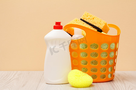塑料篮子, 带防护手套, 洗碗液瓶, 玻璃和瓷砖清洁剂, 微波炉和炉灶的洗涤剂, 米色背景上的海绵。清洗和清洁的概念.