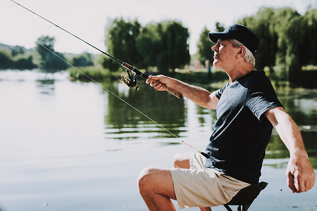 运动夏摄影照片_夏天在河上钓鱼的老人。放松的户外。坐在爷爷那里湖边的人。手里拿着钓鱼竿。夏天的运动。船长的老人。周末在河上。老年渔民.
