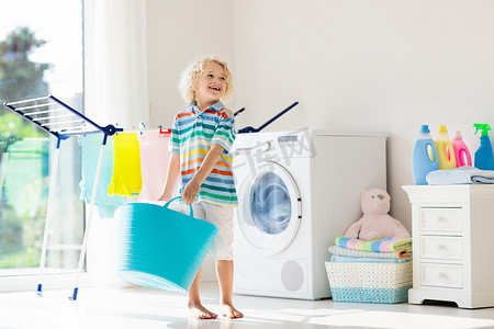 烘干机摄影照片_儿童在洗衣房内设有洗衣机或滚筒烘干机。帮助家庭琐事的孩子。现代家庭设备和洗涤洗涤剂在白色阳光的家。烘干机架清洗洗衣服. 
