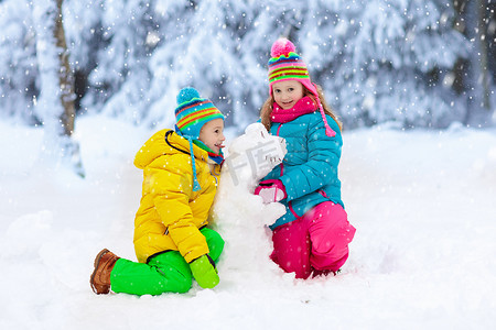 雪地里玩耍摄影照片_在雪冬公园里做雪人的孩子们。孩子们在雪地里玩耍。男孩和女孩在五颜六色的夹克和帽子修造雪恐龙在冬天庭院在雪以后。冷冬日的户外娱乐.