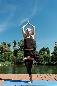 妇女在树姿势练习瑜伽 (Vrksasana) 和在公园附近的河边的瑜伽垫上做合十手势