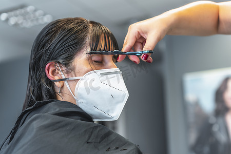 年轻的黑发女孩，戴着面罩，在美发店修剪刘海。在Covid-19大流行病中，重新开放，并为理发师采取安全措施。新的正常的，大脑皮层，社会距离
