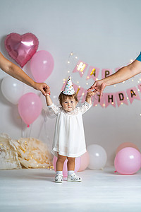 第一年的生日。身穿白衣、头戴节日帽的女婴站在背景上，戴着花环和气球，与她的父母牵着手。第一天出生。装饰生日。复制空间.