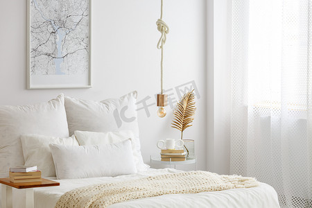 在床上挂着白色被褥、书和金色蕨叶的绳子上的简单灯泡灯在白色卧室内部的一个末端桌上