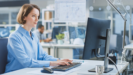 In the Industrial Engineering Facility: Portrait of the Smart and Beautiful Female Engineer working on Desktop Computer.背景专家和从事绘图和绘图工作的技术人员