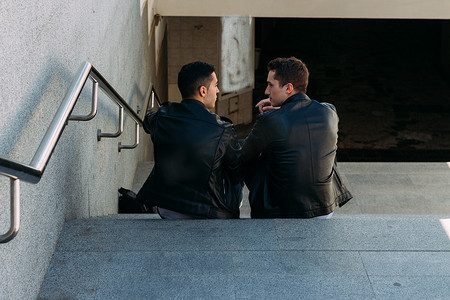 两个栏杆摄影照片_两个穿着黑色夹克、牛仔裤和白鞋的家伙正坐在台阶上。在手表的手上。社交, 结交朋友.