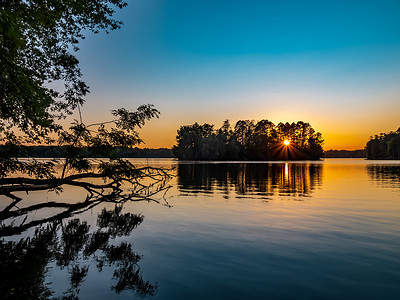 夏末在宾夕法尼亚的Pymatuning湖上落日。前景是一棵倒下的树，太阳升起来穿过背景岛上的树木，天空是橙色和蓝色的.