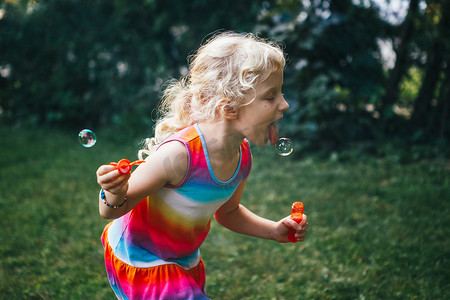 孩子试图抓住吃肥皂气泡与舌头。在夏天日落时在家后院玩的可爱滑稽的金发白种女孩的坦率的肖像。真正真实的快乐童年时刻.