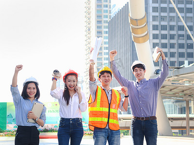 四位身穿安全背心、头戴安全帽的年轻亚洲男男女女组成的团队，站在一起举手表决，庆祝在摩天大楼背景模糊的现代城市中心实现团队合作的目标.