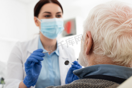 dentist in medical mask examining teeth of senior man with probe and mirror in dental chair