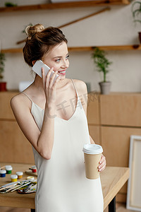 美丽微笑的年轻妇女拿着咖啡去和谈话的智能手机在艺术工作室