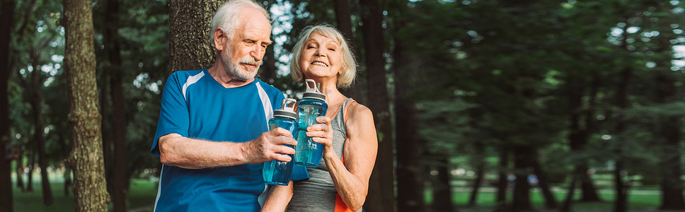 在公园里，一个微笑的老年妇女在丈夫身边拿着运动瓶的全景照片 