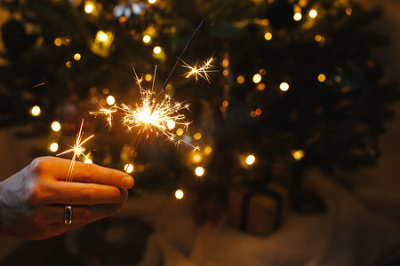 手拿着烟火，照亮着装饰华丽的树。新年快乐！在黑暗的房间里，男人手拿着点燃的火花，背景是圣诞树上的灯。大气庆祝活动