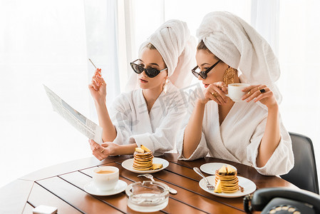 穿着浴袍、太阳镜和珠宝的时尚女性，头上有毛巾，一边抽烟一边看报纸，一边吃早餐 