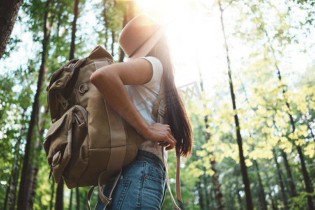 后面的旅行背包和时髦的女孩戴着帽子的看法。年轻勇敢的妇女独自旅行在森林中的树木在户外。镜头光晕效果